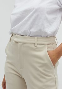 Pantaloni beige su misura con una texture liscia, dotati di tasche laterali e una vestibilità frontale piatta. Indossati con una camicia bianca morbida.