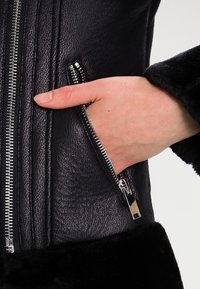 Svart läderjacka med en ficka med blixtlås, texturerad yta och fleece-fodrade ärmar, som framhäver silverfärgad hårdvara och en kroppsnära design.