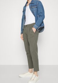 Veste en denim avec deux poches poitrine, débardeur blanc, pantalon plissé vert olive et baskets beiges avec œillets multicolores.