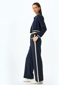 Marineblauw trainingspak met een cropped jacket voorzien van witte en groene strepen en een wijde broek met een beige zijstreep; glad, gestructureerd materiaal.