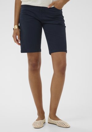 Nő tengerészkék térdig érő rövidnadrágot, krémszínű kötött felsőt, arany karkötőt és krémszínű fonott papucsot visel, egyik kezét a zsebében tartva áll.