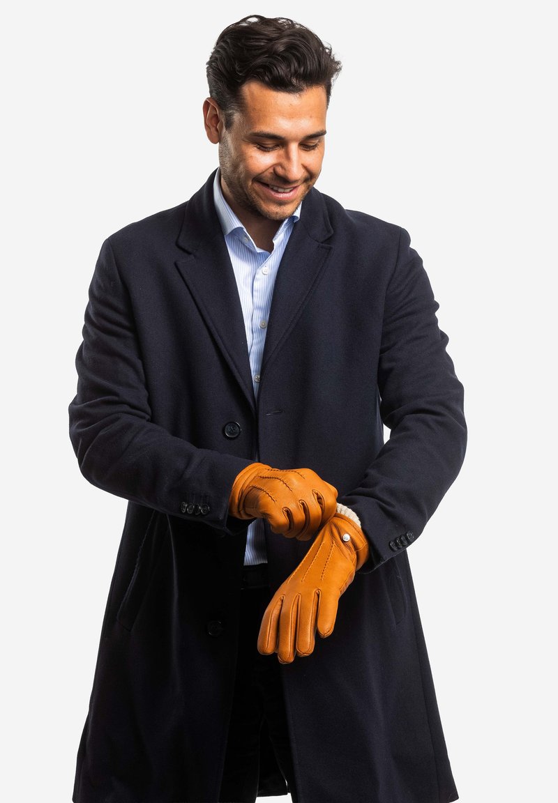 Leather gloves in tan, featuring a fitted design and stitching details. Worn by a person in a navy coat, showcasing a textured finish.