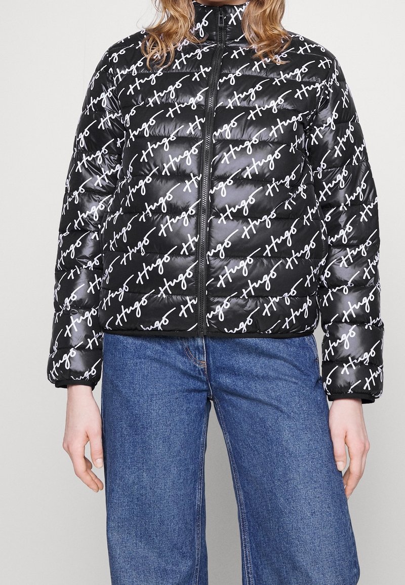 Sort pufferjakke med et gentagende hvidt tekstmønster, høj krave, frontlynlås og quiltet tekstur. Kombineret med blå denimjeans.