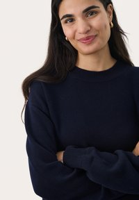 Pull en tricot bleu marine avec un col rond côtelé et des manches longues légèrement bouffantes. Texture douce, porté de manière décontractée avec les bras croisés.