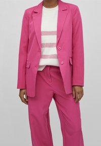 Rosa kavaj med två knappar och fickor, tillsammans med en vit tröja med rosa ränder; vida rosa byxor fulländar outfiten.