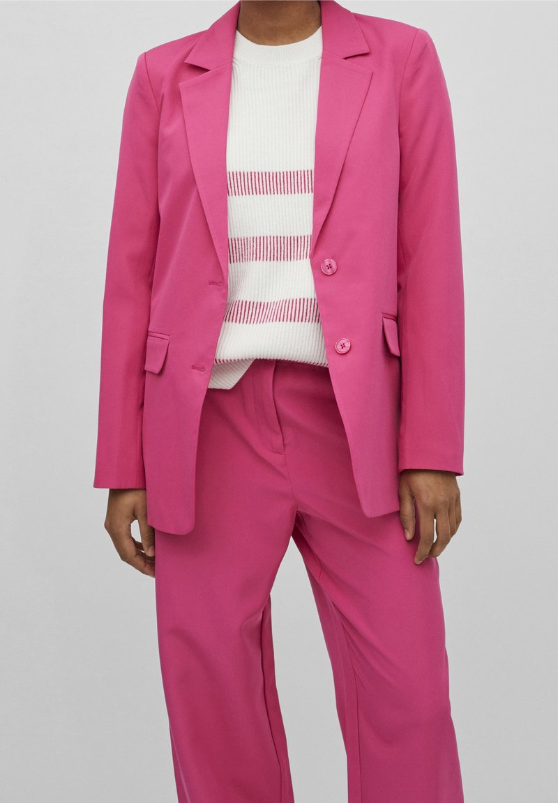 Rosa kavaj med två knappar och fickor, tillsammans med en vit tröja med rosa ränder; vida rosa byxor fulländar outfiten.