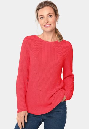 Pull en tricot corail à manches longues, col rond, texture côtelée et détails de couture diagonale. Porté avec un jean bleu.