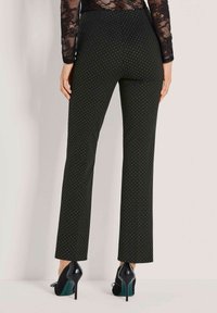 Vrouw in zwarte hoge taille broek met kleine witte stippen, zwarte kanten top en zwarte hoge hakken, staand tegen een lichte muur.