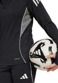 Zwarte sporttop met witte accenten en logo, gecombineerd met zwarte broek. Houdt een zwart-wit gestreepte voetbal vast.