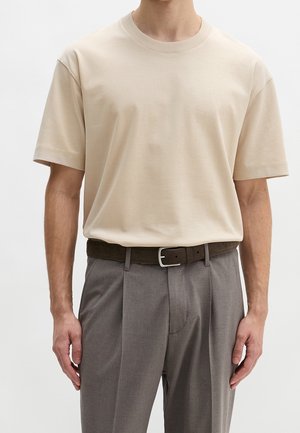 Hombre con una camiseta beige de manga corta y cuello redondo metida dentro de unos pantalones grises plisados con un cinturón marrón oscuro.