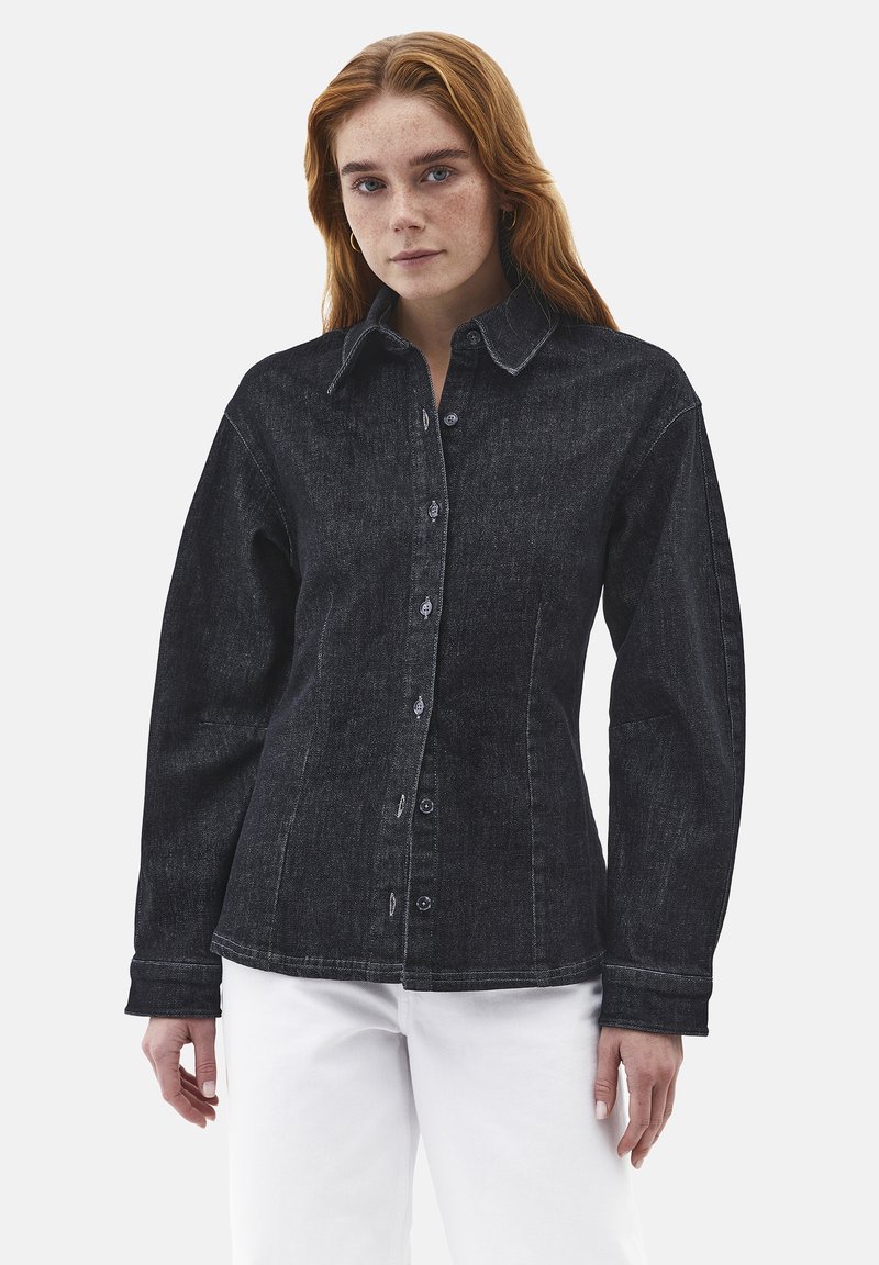 Kvinde med rødt hår iført en tætsiddende sort denimjakke og hvide bukser, stående mod en ensfarvet lys baggrund.
