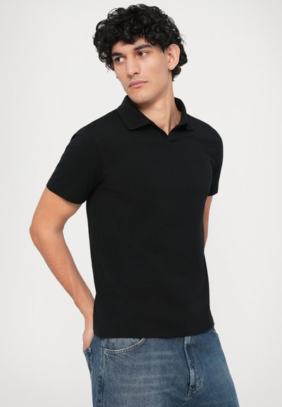 Polo nero in cotone con colletto classico e maniche corte, caratterizzato da un design aderente e una texture liscia. Indossato con jeans blu.