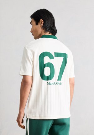 Uomo con capelli neri che indossa una camicia bianca a maniche corte con colletto verde, grande "67" verde e "Marc O'Polo" sulla schiena, pantaloni verdi con strisce bianche.