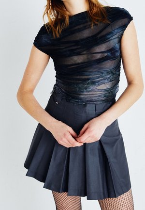 Woman wearing a black sheer, layered top, a pleated dark skirt, and fishnet stockings, standing with hands clasped in front on a white background.