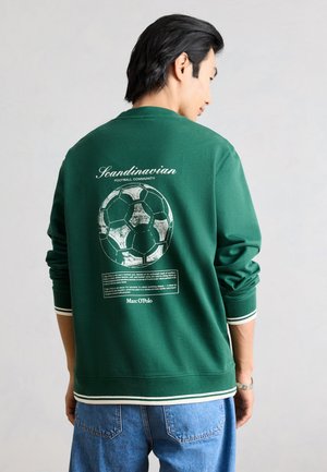 Uomo che indossa una felpa verde con grafica di pallone da calcio bianco e scritta "Scandinavian Football Community", abbinata a jeans blu.