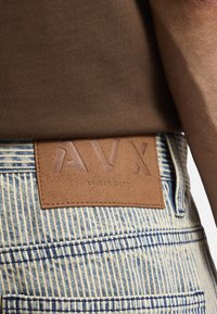 Camisa de algodón marrón acanalada sobre un pantalón de mezclilla a rayas azules y blancas, con un parche de cuero marrón que muestra "AVX" grabado y "AMIREY DEPT." debajo.