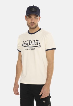 T-shirt crème à manches courtes avec col côtelé marine, arborant le logo "Von Dutch CALIFORNIA" en bleu foncé. Shorts cargo noirs. Casquette marine.