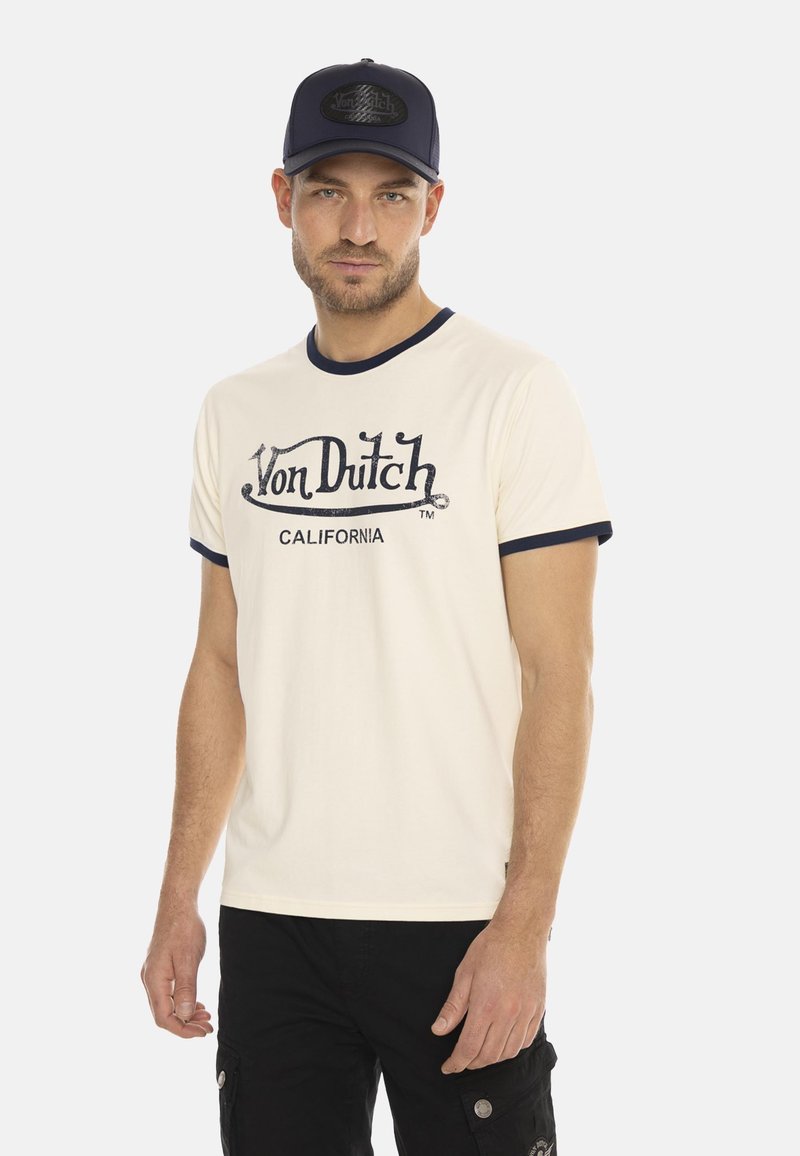 T-shirt crème à manches courtes avec col côtelé marine, arborant le logo "Von Dutch CALIFORNIA" en bleu foncé. Shorts cargo noirs. Casquette marine.