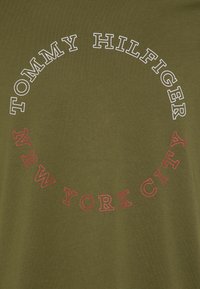 Camiseta de algodón verde oliva con texto blanco y rojo. El texto, dispuesto en un diseño circular, dice "TOMMY HILFIGER NEW YORK CITY."