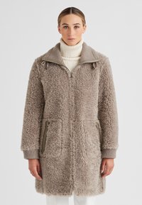 Frau in einem taupefarbenen Sherpa-Fleece-Mantel mit Reißverschlusstaschen über einem cremefarbenen Rollkragenpullover, die vor einem schlichten Hintergrund steht.