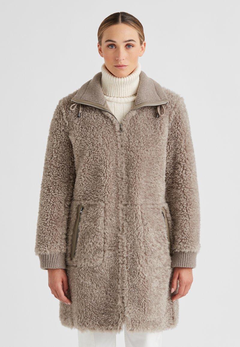 Frau in einem taupefarbenen Sherpa-Fleece-Mantel mit Reißverschlusstaschen über einem cremefarbenen Rollkragenpullover, die vor einem schlichten Hintergrund steht.