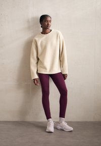 Felpa oversized in crema e texture abbinata a leggings bordeaux; sneakers rosa pallido e calzini alla caviglia visibili completano l'outfit.