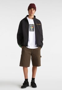 Zwarte bomberjack met groene accenten, wit T-shirt met een grafische afbeelding, bruine shorts en zwarte sneakers. Donkere beanie op het hoofd.