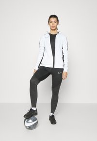 Nike sportoutfit met een witte zip-up jas met zwarte accenten, zwarte strakke leggings met een textuurpatroon en zwarte voetbalschoenen.