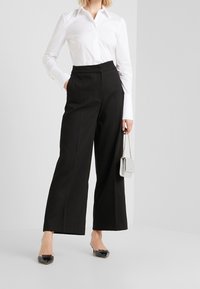 Eine Frau trägt ein weißes, gebuttontes Hemd und hochgeschnittene schwarze weite Hosen und hält eine kleine silberne Kettenhandtasche.