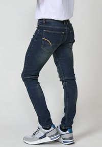 Jeans de mezclilla oscuros de corte ajustado con costuras amarillas, dos bolsillos traseros y cinturilla elástica, combinados con zapatillas grises con acentos azules.