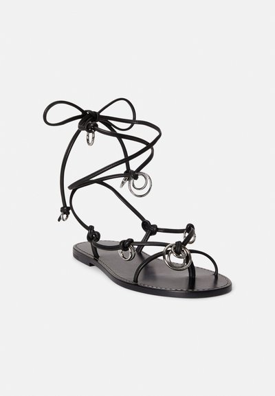 Μαύρο δερμάτινο σανδάλι με λεπτές τιράντες και κυκλικές μεταλλικές διακοσμήσεις. Η επίπεδη σόλα έχει σχεδιασμό με στρώσεις και φιόγκο δεμένο στον αστράγαλο.