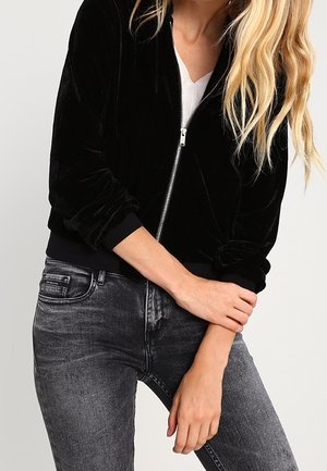 Frau trägt eine schwarze Samtjacke mit Reißverschluss über einem weißen Hemd und ausgewaschenen schwarzen Jeans, mit langem blondem Haar sichtbar.