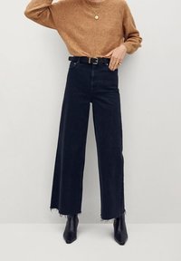 Jeans larghi neri con orli sfrangiati, indossati con un maglione lavorato marrone e stivali neri con tacco. Caratterizzati da una cintura nera con fibbia dorata.