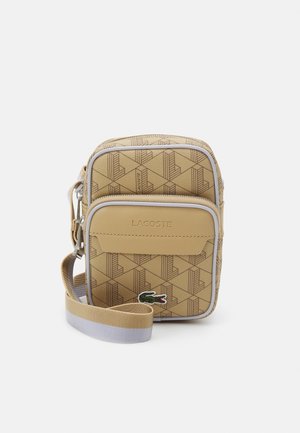 Pequeño bolso cruzado beige con patrón geométrico, bolsillo frontal con cremallera, logotipo de Lacoste y correa ajustable sobre fondo blanco.