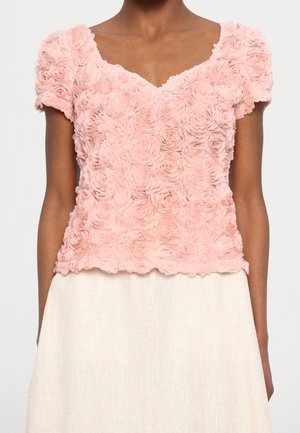 Femme portant un chemisier rose clair à manches courtes avec texture florale 3D et jupe crème, debout devant un fond uni.
