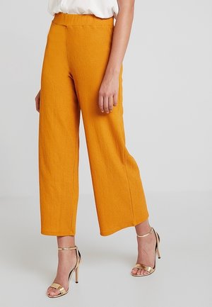 Femme portant un pantalon large texturé couleur moutarde coupé au-dessus de la cheville, associé à une blouse blanche et des sandales à talons hauts dorées à brides, sur un fond uni.