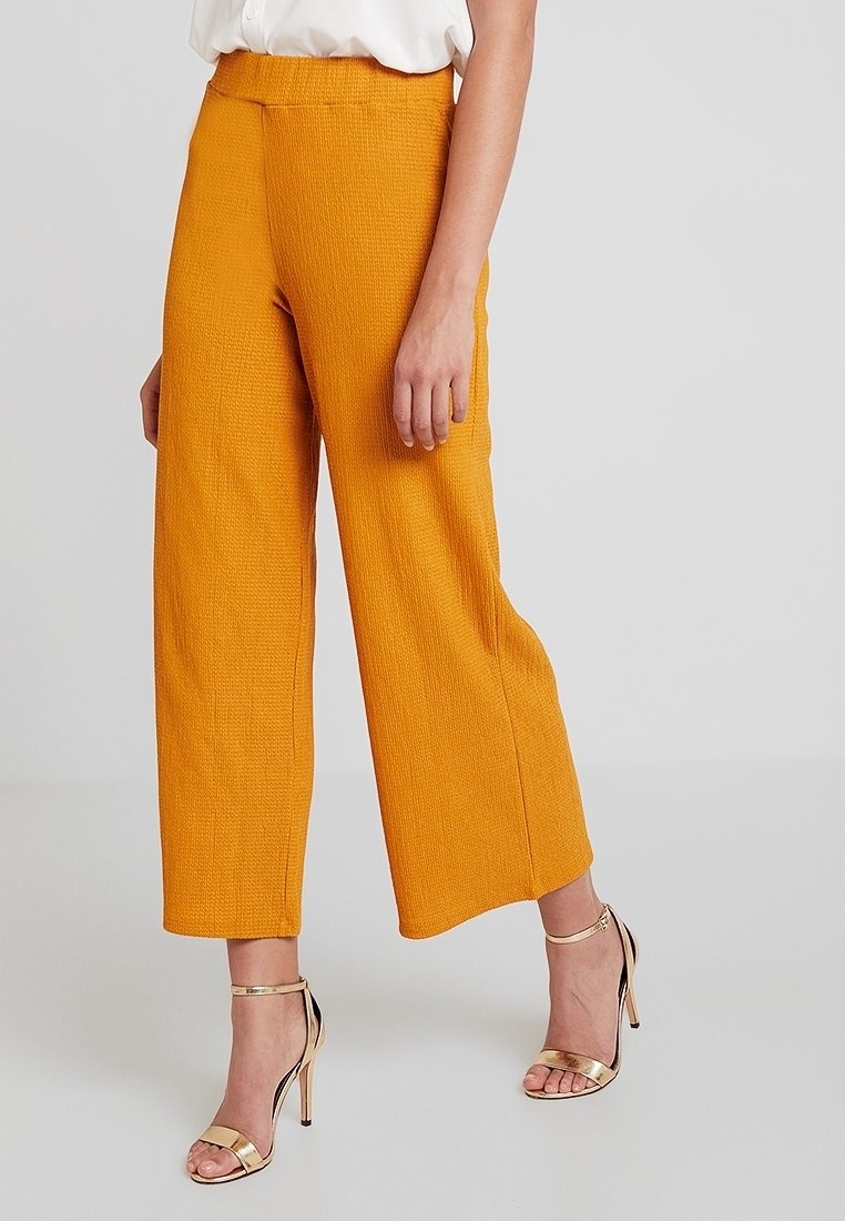 Femme portant un pantalon large texturé couleur moutarde coupé au-dessus de la cheville, associé à une blouse blanche et des sandales à talons hauts dorées à brides, sur un fond uni.