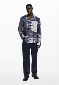 Long-sleeve button-up shirt featuring a patchwork design in shades of blue and white, with various patterns and textures. Paired with dark pants.