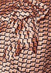 Tissu soyeux avec motif carré géométrique aux nuances de rose, marron et beige, comportant un nœud noué et un bouton noir visible.