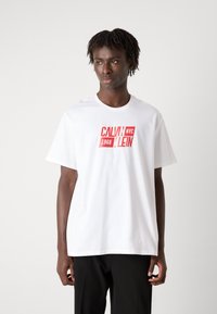 Bela bombažna majica s kratkimi rokavi, z rdečim grafičnim tiskom na sredini, ki prikazuje napis "CALVIN KLEIN NYC 1968".