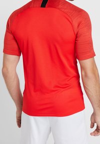 Hombre con una camiseta atlética roja de manga corta y pantalones cortos blancos, visto desde atrás sobre un fondo simple.
