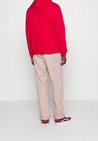 Sudadera de algodón roja, pantalones beige, zapatillas de deporte burdeos. La sudadera tiene una gran capucha y puños acanalados; los pantalones tienen un corte relajado con dobladillos enrollados.