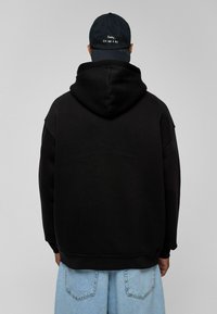 Zwarte oversized hoodie met een capuchon, gemaakt van fleece-materiaal. Gecombineerd met lichtblauwe wijde jeans. De pet heeft witte tekst: "Sorry, ik ben geen DJ."