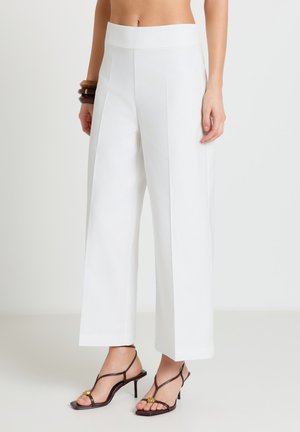 Pantaloni bianchi a vita alta e gamba larga indossati con sandali con tacco scuri color marrone con cinturini e braccialetti di legno su un polso.