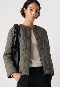 Gequiltde olijfgroene jas met drukknoopjes, ronde halslijn en textuurstiksel. Gemaakt met een beige gebreid top en zwarte broeken.