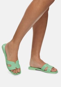 Sandales vertes avec un matériau lisse, une conception ouverte au niveau des orteils, des brides crantées et une semelle plate avec une silhouette simple.