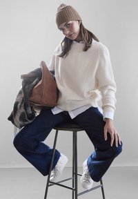 Suéter de punto ribeteado crema, camisa blanca con cuello debajo, pantalones vaqueros oscuros de pierna ancha, bolso de ante marrón, chal gris con estampado, gorro de punto beige, zapatillas blancas.