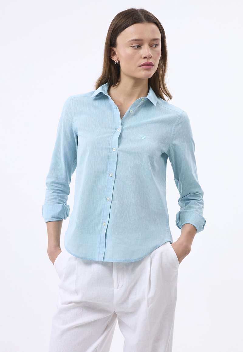 Camisa de botones azul claro con una textura sutil, mangas largas remangadas en los puños y un pequeño logo bordado en el pecho.