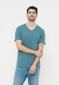 Άνδρας με κοντά σκούρα μαλλιά που φοράει τιρκουάζ μπλουζάκι V-neck και μπλε τζιν, στέκεται μπροστά σε απλό φωτεινό φόντο, κρατώντας τον αριστερό του πήχη.