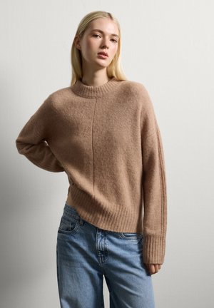 Jonge vrouw met lang blond haar, gekleed in een lichtbruine gebreide trui en blauwe jeans, staand met één hand op haar heup tegen een effen muur.